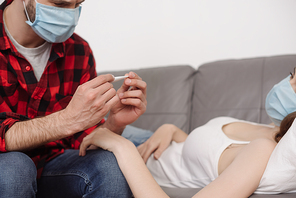 cropped view of man in medical mask looking at thermometer near sick girlfriend lying on sofa