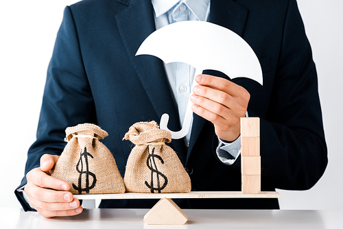 cropped view of businessman holding carton umbrella near scales with money bags and wooden blocks isolated on white