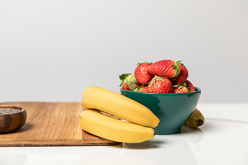 sweet and tasty strawberries in bowl near yellow bananas and cutting board on grey