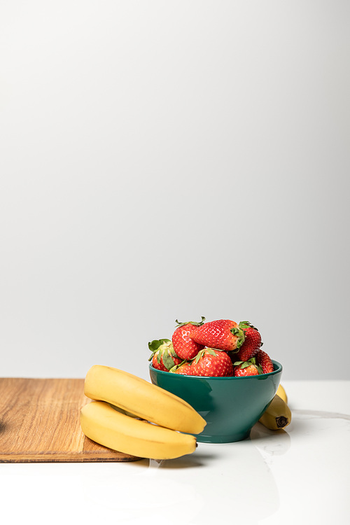 sweet strawberries in bowl near yellow bananas and cutting board on grey