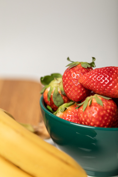 selective focus of red strawberries in bowl near yellow bananas on grey