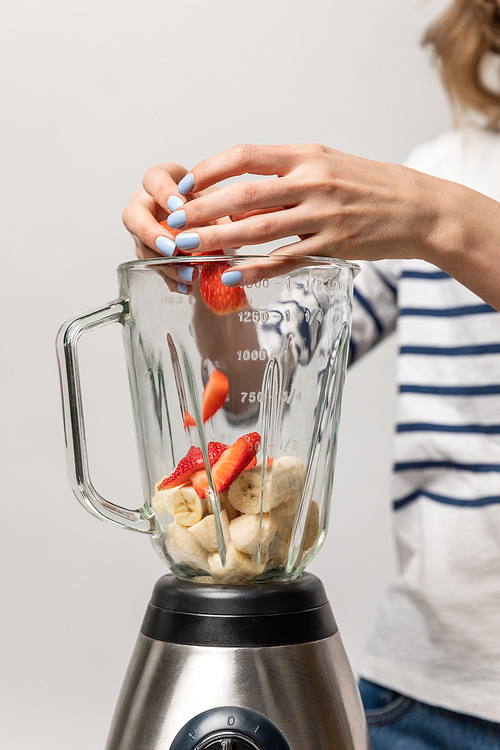 cropped view of woman putting red strawberries in blender with bananas on white