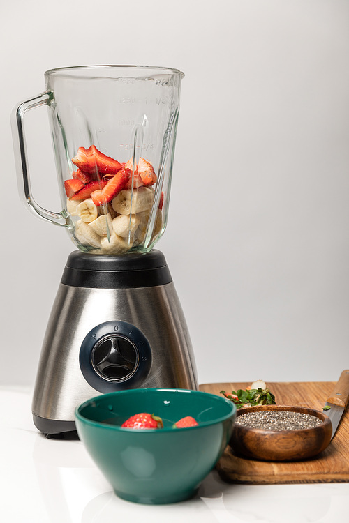 sweet strawberries and ripe bananas in blender near bowls and cutting board on grey