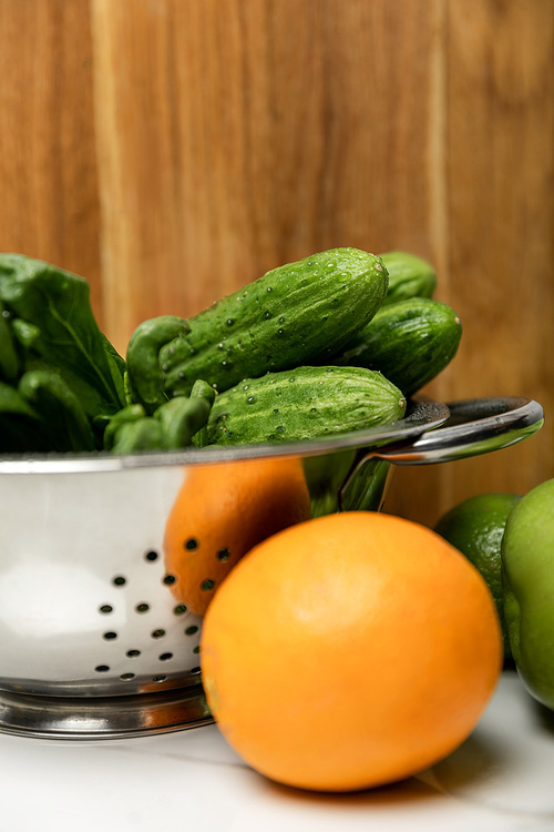selective focus of green cucumbers in colander near orange and wooden cutting board