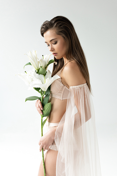 tender young woman in beige lingerie and mesh sleeves looking at lilies isolated on white