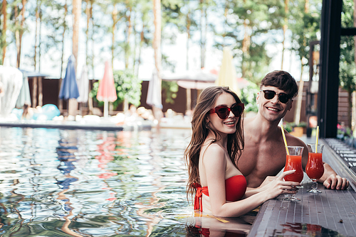happy young couple relaxing in swimming pool with glases of refreshing drink an smiling at camera