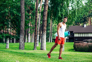 cheerful bearded man holding fitness mat while walking in park