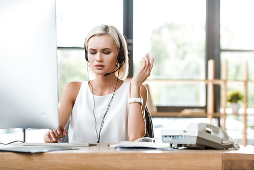 selective focus of upset blonde woman in headset gesturing while working in office