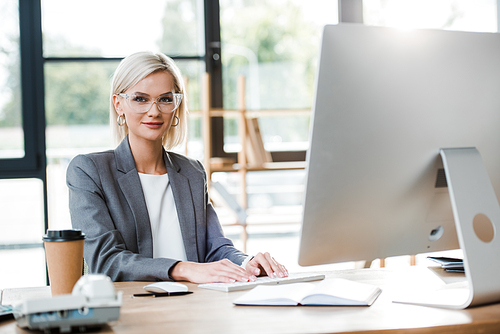 selective focus of happy businesswoman in glasses working in modern office near paper cup