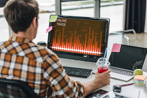 back view of young programmer holding glass of juice while sitting at workplace near computer monitor with online trade on screen