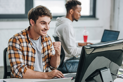 cheerful programmer showing winner gesture while working near african american colleague