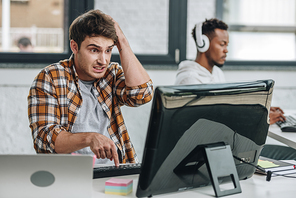 selective focus of discouraged programmer holding hand on head while looking at monitor near african american colleague