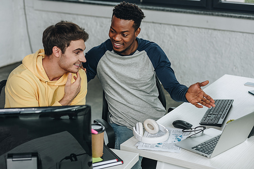 cheerful african american programmer pointing at computer monitor while sitting near colleague