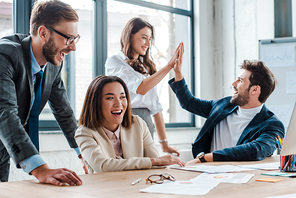 selective focus of happy businessman in glasses smiling near multicultural coworkers giving high five in office