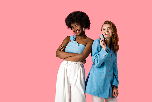 happy redhead girl holding credit card near african american woman standing with crossed arms isolated on pink