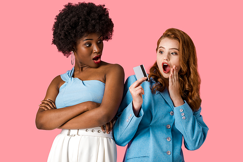 surprised redhead girl holding credit card near shocked african american woman standing with crossed arms isolated on pink