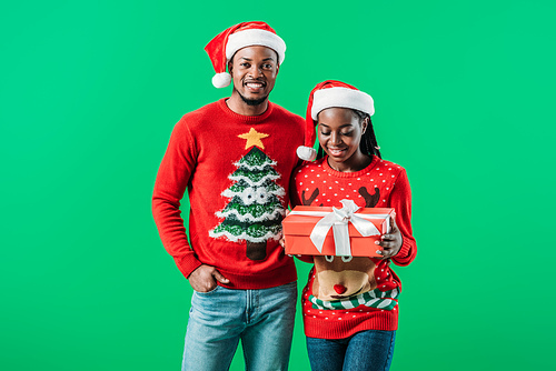 African American man in red Christmas sweater and Santa hat near woman holding gift box isolated in hands on green