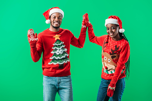 African American couple in red Christmas sweaters and Santa hats giving high five to each other and  isolated on green