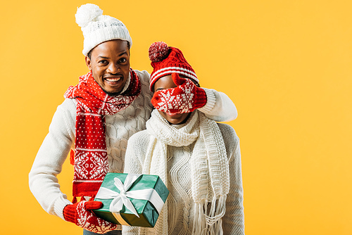African American man in cozy winter outfit holding green gift box and covering with hand eyes of woman isolated on yellow