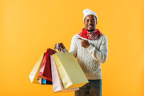 African American man in winter outfit holding shopping bags and pointing with finger isolated on yellow