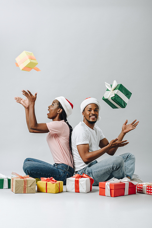 African American couple in Santa hat sitting back to back and throwing up gift boxes isolated on grey