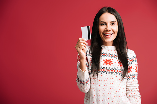 happy young woman in winter sweater holding credit card, isolated on red