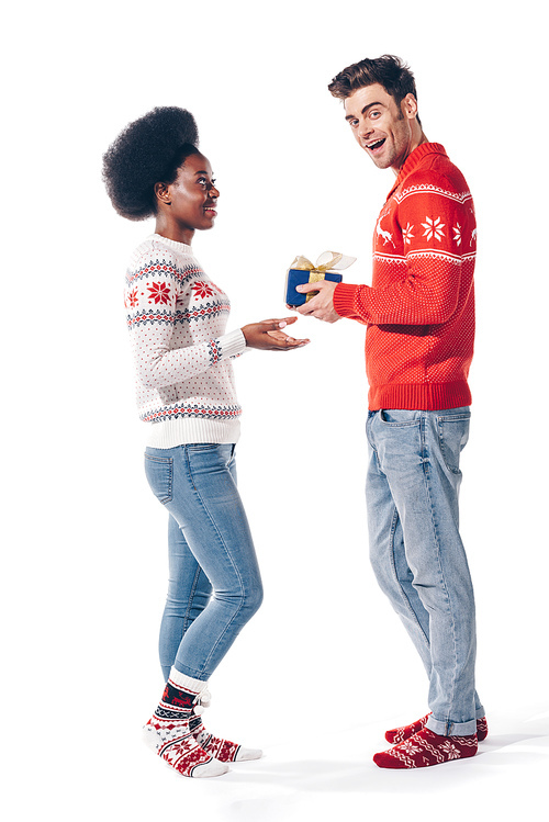 smiling interracial couple in winter sweaters holding christmas present, isolated on white