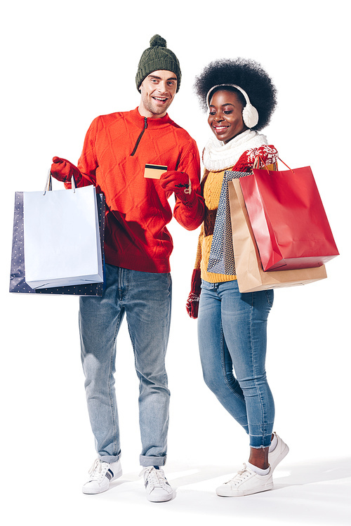 happy multiethnic couple in winter sweaters holding credit card and shopping bags, isolated on white