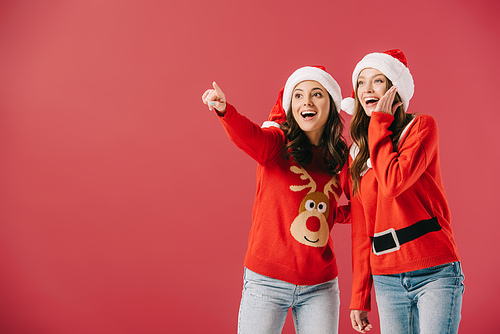 attractive and smiling women in sweaters and santa hats pointing with finger isolated on red