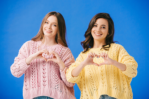attractive and smiling women in sweaters showing heart gesture isolated on blue