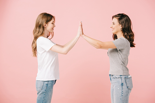 side view of attractive and smiling women in t-shirts giving high five on pink background