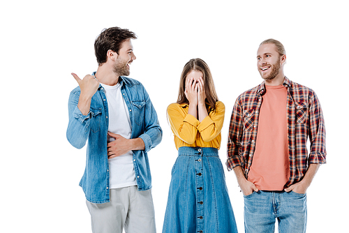 young man pointing with finger away near smiling friend and girl covering face with hands isolated on white