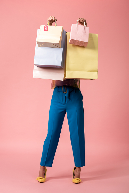 full length view of stylish disco girl holding shopping bags in front of face on pink