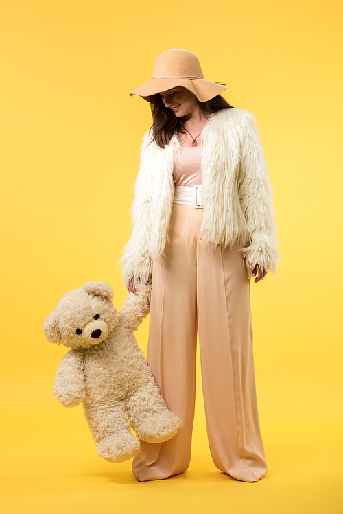 happy elegant girl in faux fur jacket and hat looking at teddy bear on yellow