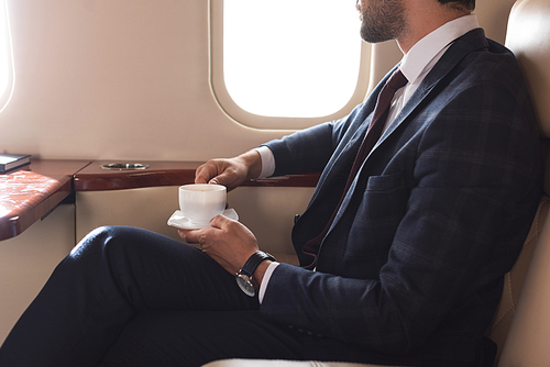 cropped view of businessman in suit holding cup of coffee in airplane during business trip