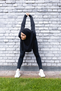 young muslim sportswoman exercising near brick wall and green grass