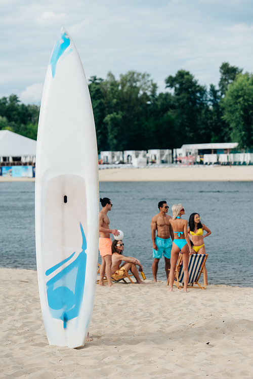 surfing board on sand near multicultural friends resting on riverside