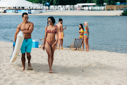 handsome mixed race man holding surf board while walking near african american girl in swimsuit