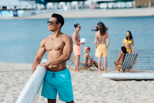 handsome mixed race man holding surfing board while multicultural friends resting near river on beach