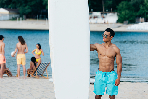 smiling, handsome mixed race man standing near surfing board on beach