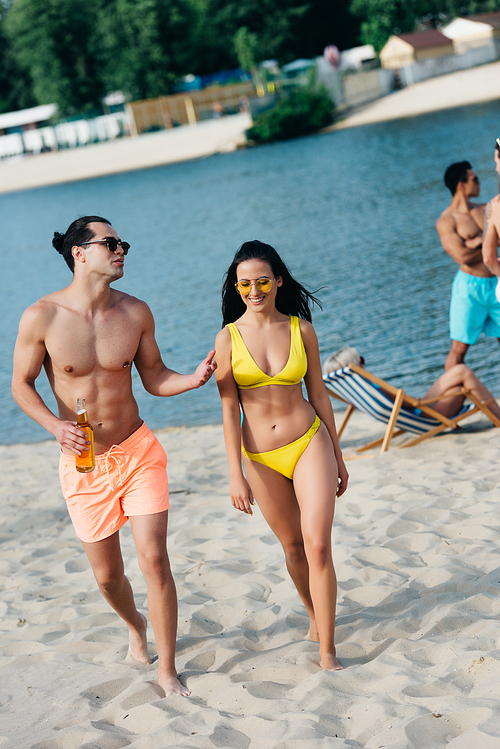 cheerful young man with bottle of beer talking to attractive girl in swimsuit while walking together on beach