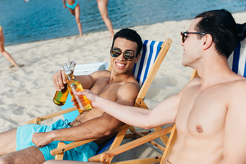 cheerful multicultural friends clinking bottles of beer while sitting in chaise lounges on beach