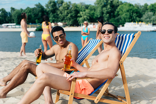 cheerful multicultural friends smiling at camera while sitting on beach in chaise lounges and holding bottles with beer
