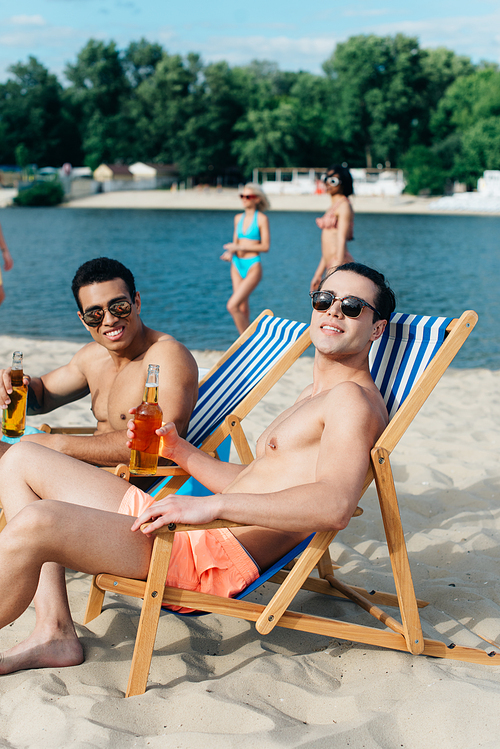 smiling multicultural friends  while sitting on beach in chaise lounges and holding bottles with beer