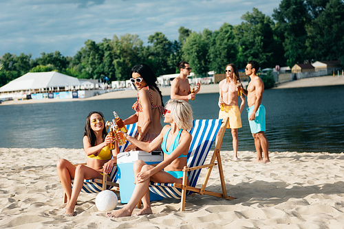 attractive multicultural girls clinking bottles of beer while resting on beach near young men