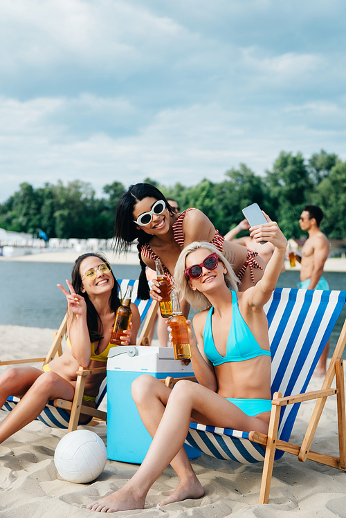 beautiful multicultural girls in swimsuits taking selfie while drinking beer on beach
