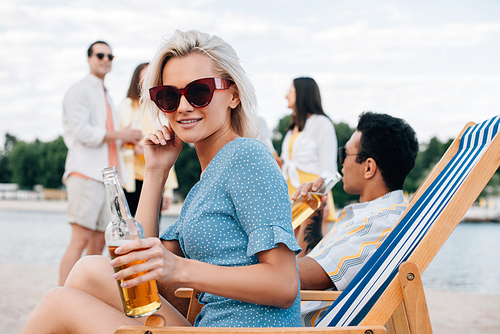 beautiful woman in sunglasses holding bottle of beer while sitting in chaise lounge near multicultural friends