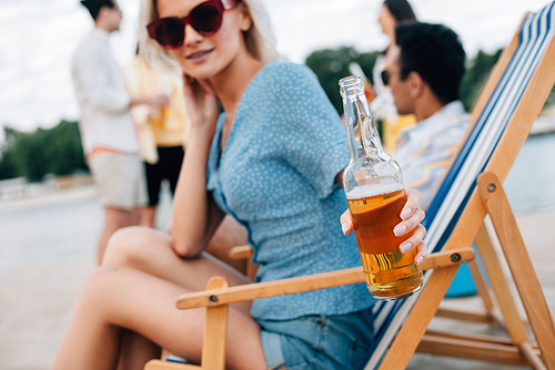 selective focus of attractive young woman in sunglasses holding bottle of beer while sitting in chaise lounge near multicultural friends