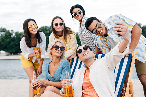 handsome young man taking selfie with multicultural friends having fun and drinking beer on beach