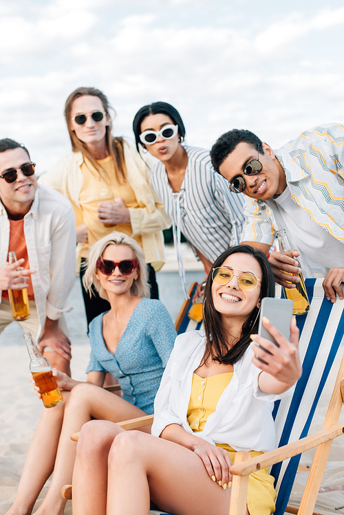 cheerful multicultural friends taking selfie while drinking beer and having fun on beach